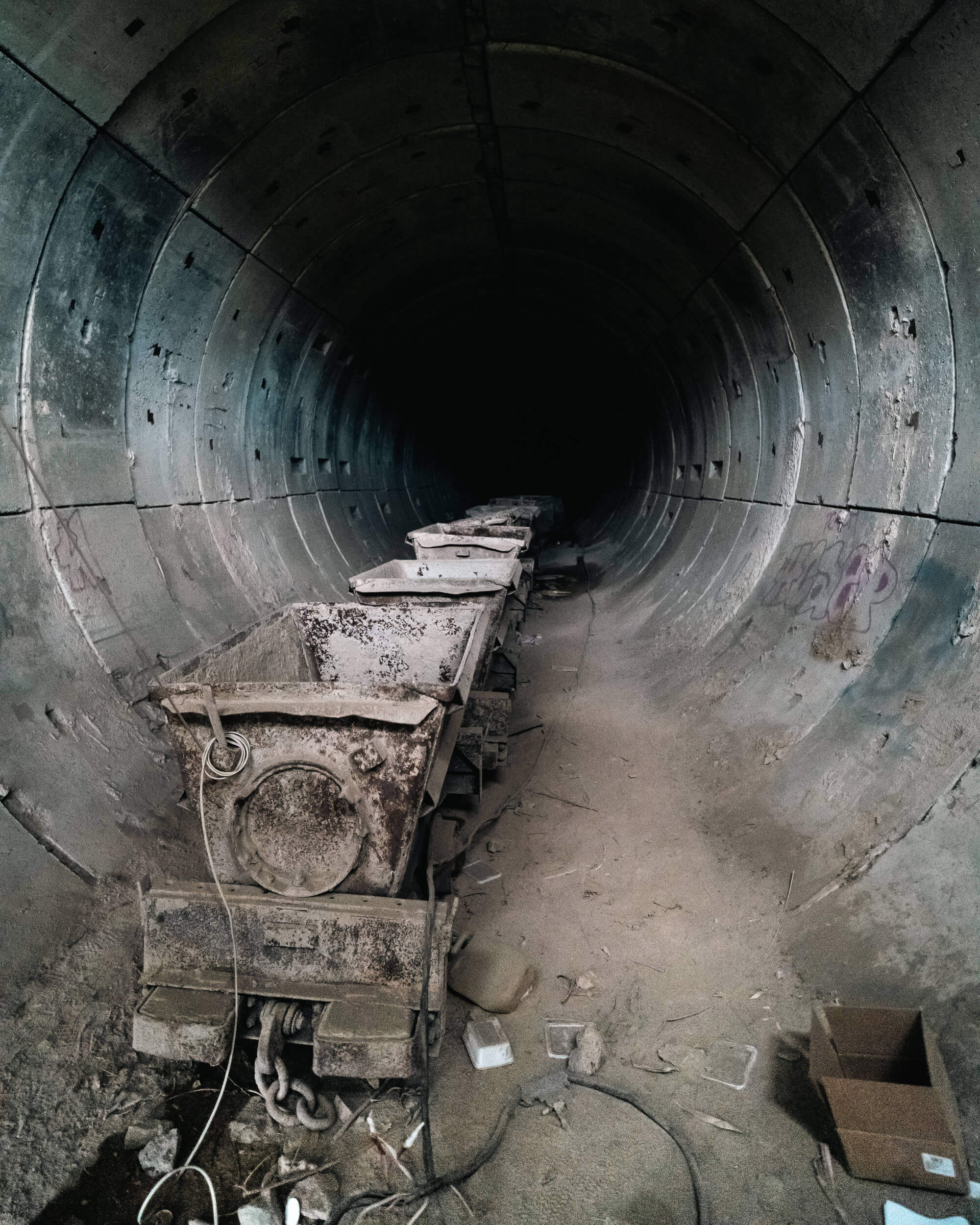 A dark tunnel with cylindrical walls, featuring dusty and rusted mining carts lined up on the ground. The far end of the tunnel fades into complete darkness, surrounded by debris and remnants of wear.