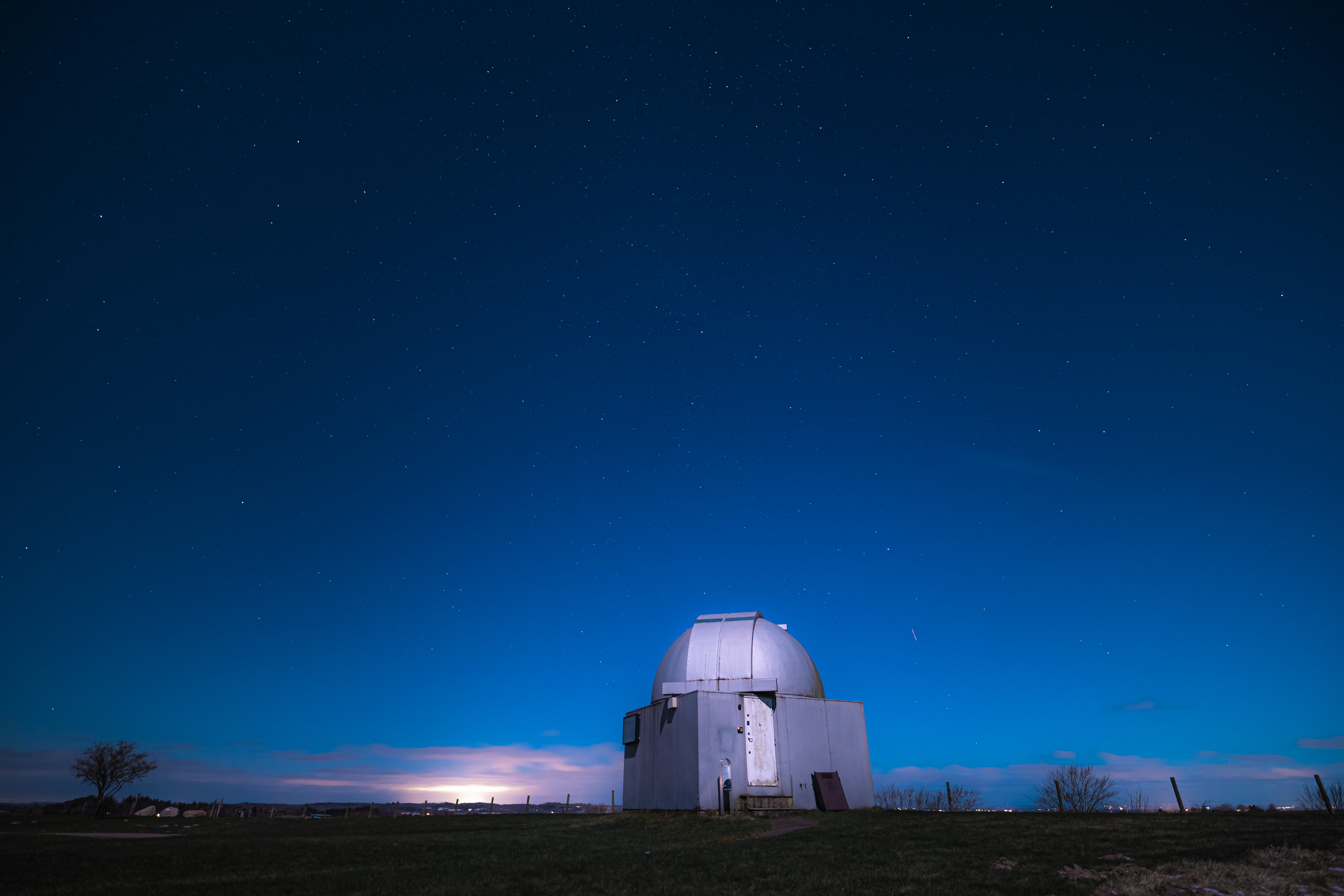 A metallic observatory dome under a starry night sky with a soft blue atmosphere, accompanied by faint distant lights on the horizon, set in a grassy landscape.