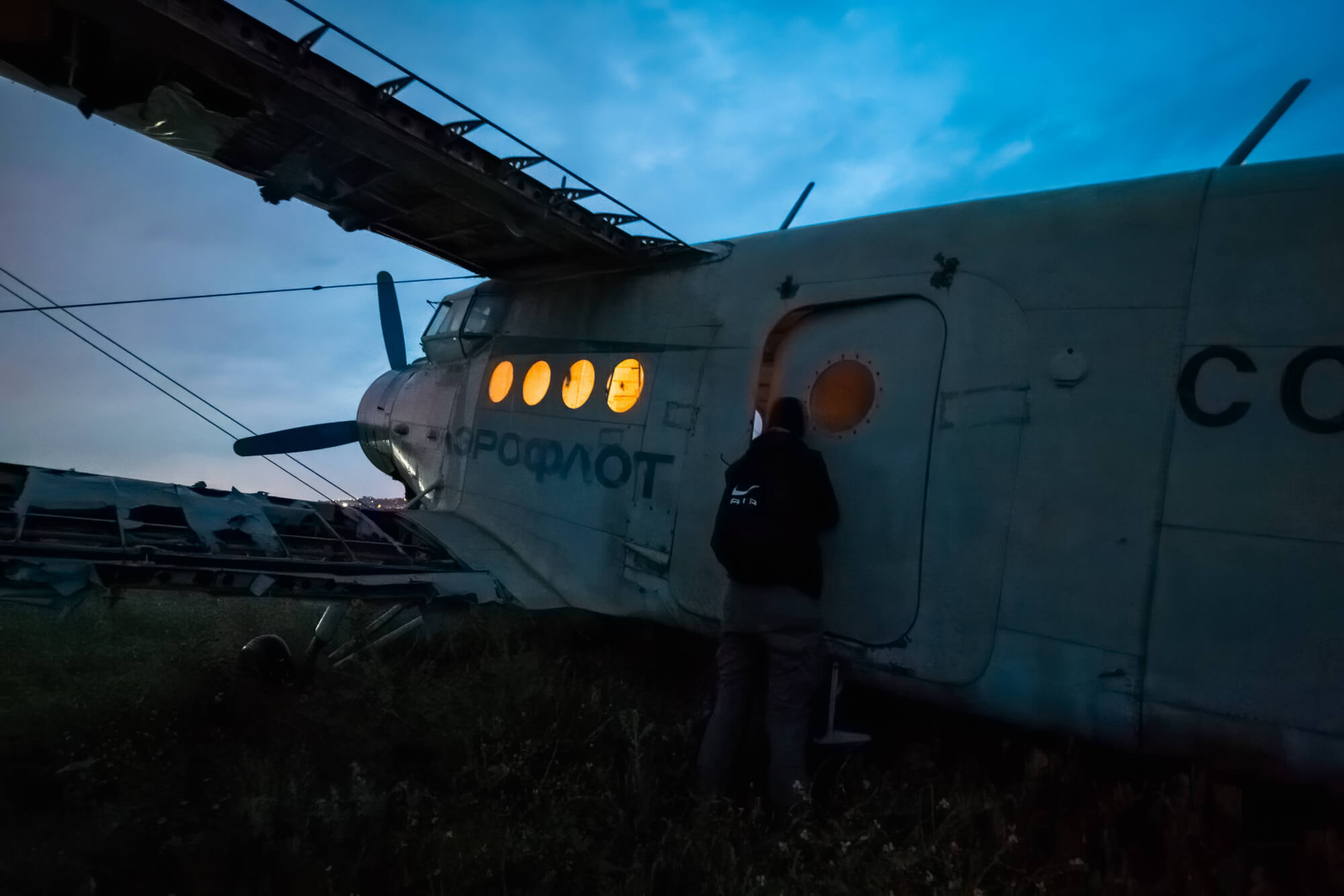 A person stands in front of an abandoned aircraft at twilight, with the plane's windows glowing orange, under a blue-grey sky.