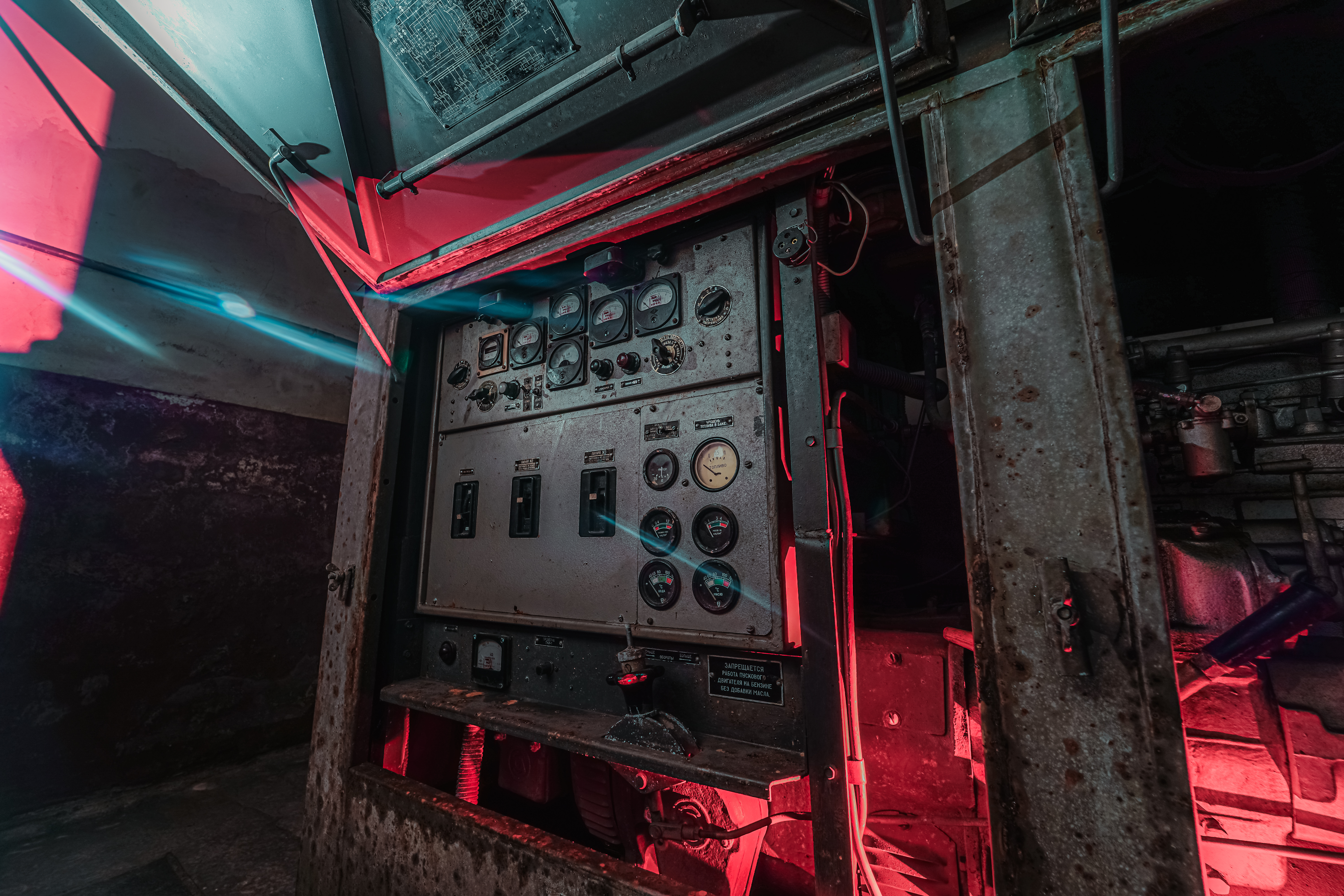 A close-up view of an industrial control panel with various gauges and switches, illuminated by red and blue light. The panel is situated in a dimly lit environment with visible textures of weathered metal and walls.