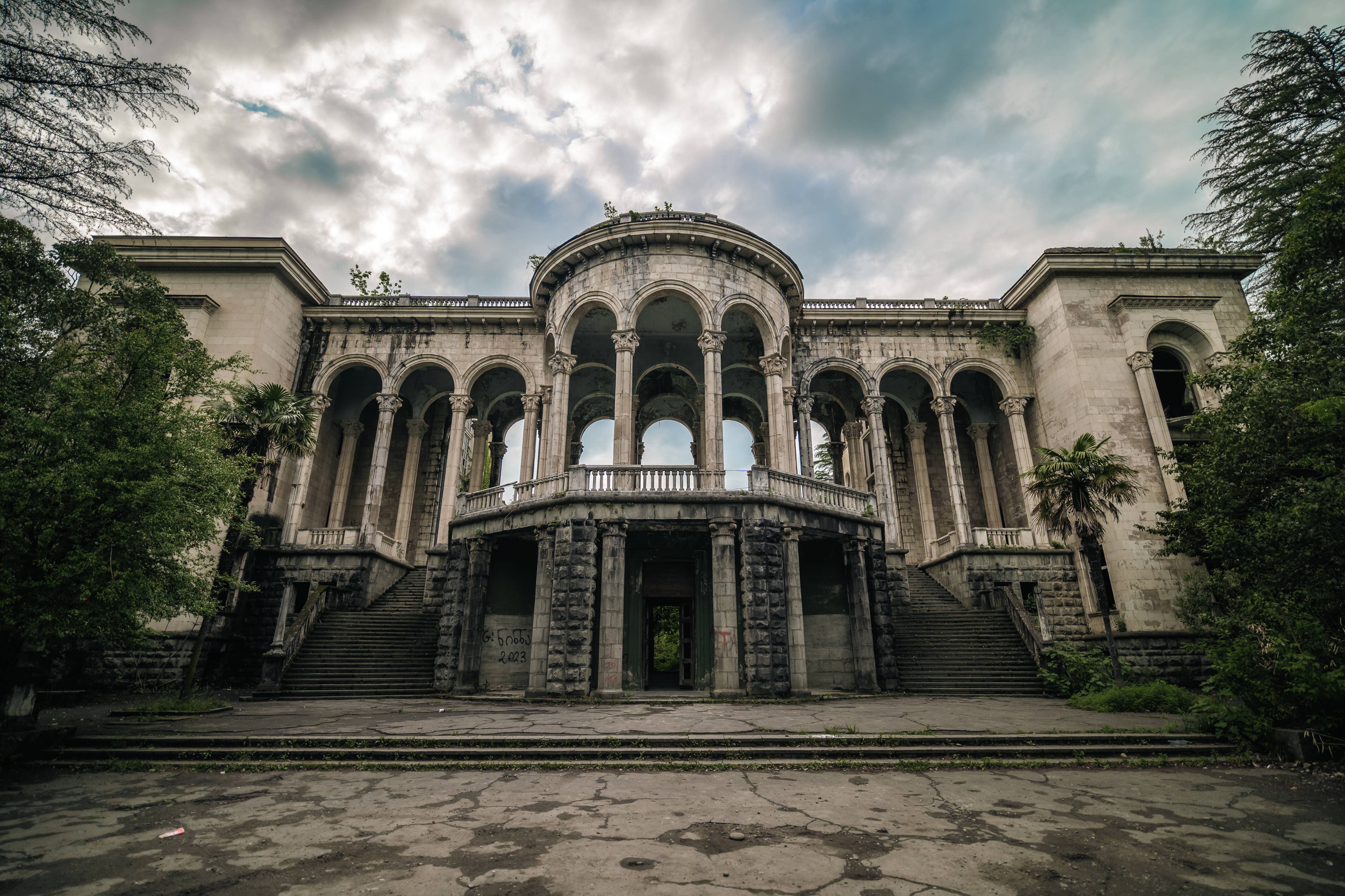 An abandoned building with grand arches and a staircase leading to the entrance, surrounded by overgrown vegetation and under a cloudy sky.