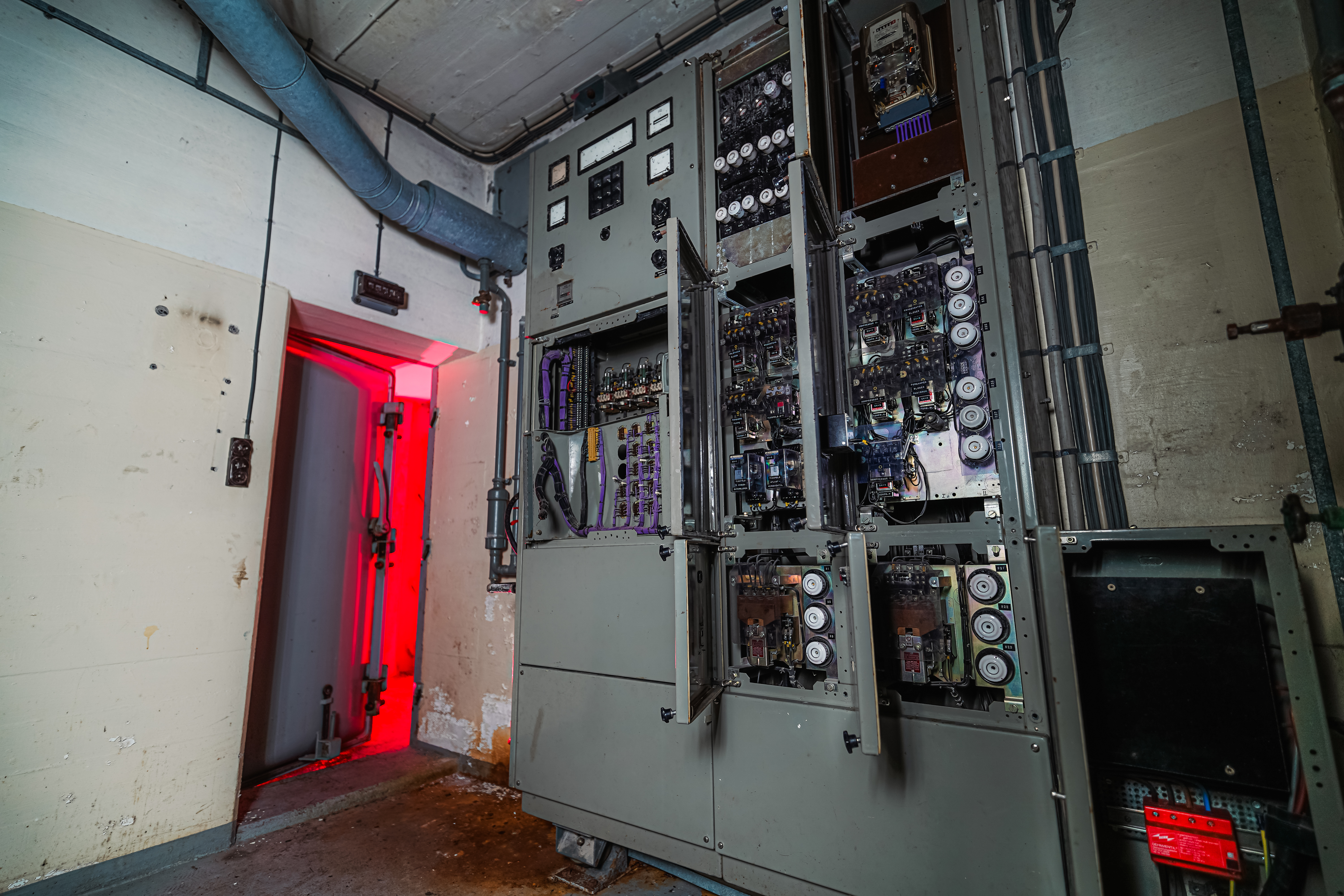 A dimly lit room featuring a large control panel with open compartments filled with machinery. A doorway on the left emits a red glow, casting light on the nearby wall, which shows signs of wear.