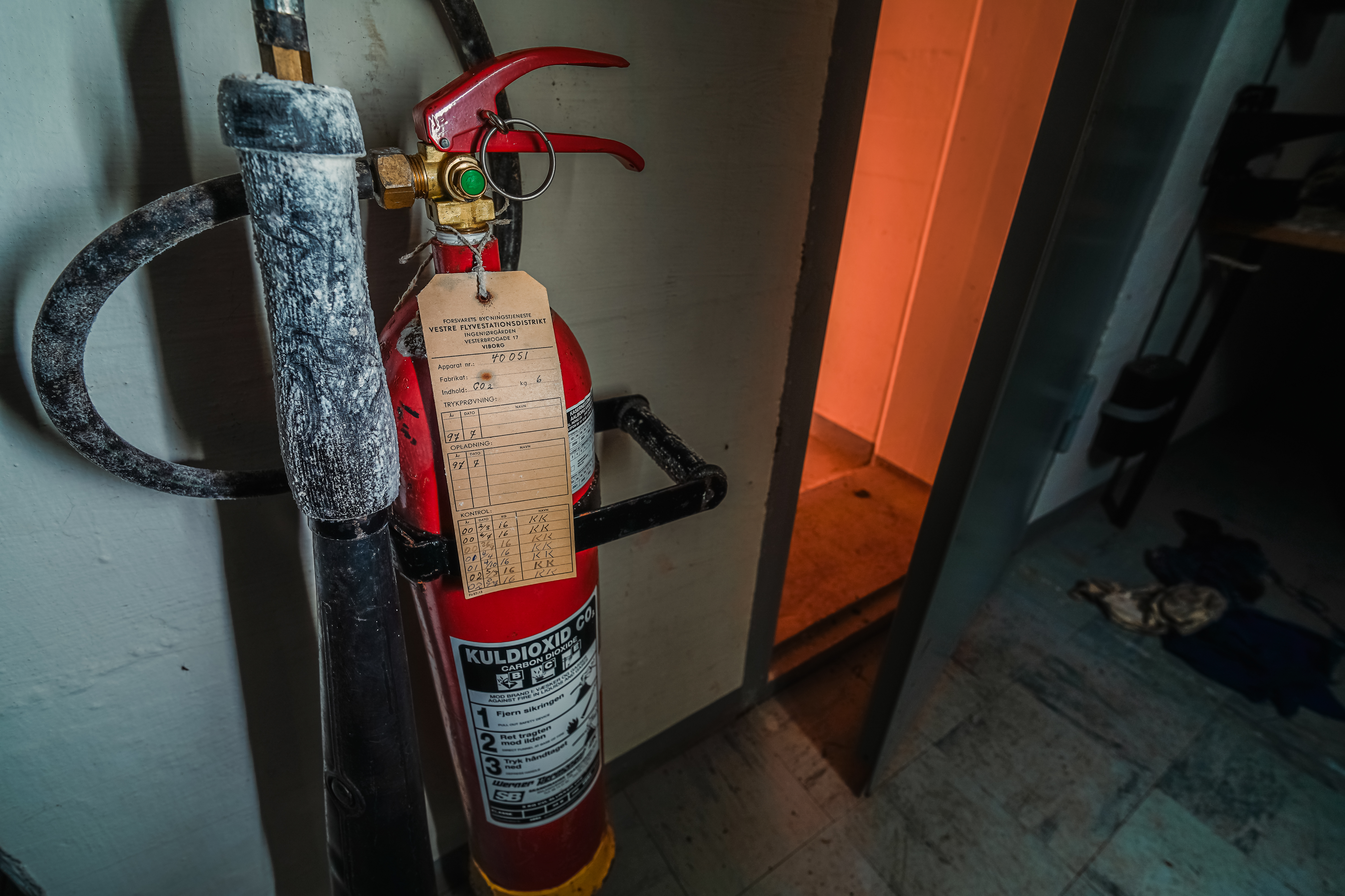 A close-up of a fire extinguisher on a wall with a service tag, illuminated by warm orange light from a doorway in the background, contrasting with the dim surroundings.