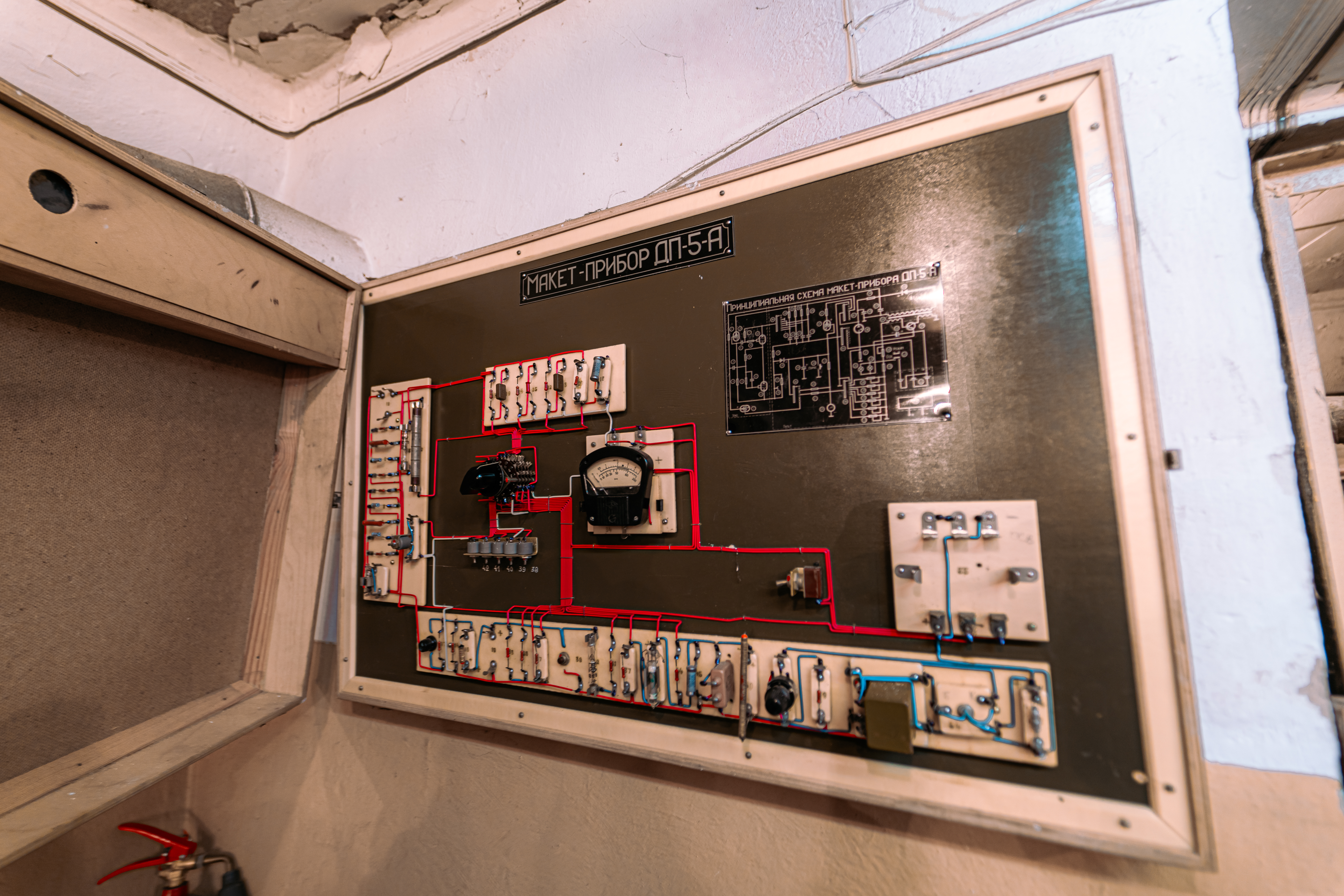 Close-up of an old electrical circuit panel with red and blue wires, components, and an analog meter, mounted on a damaged wall.