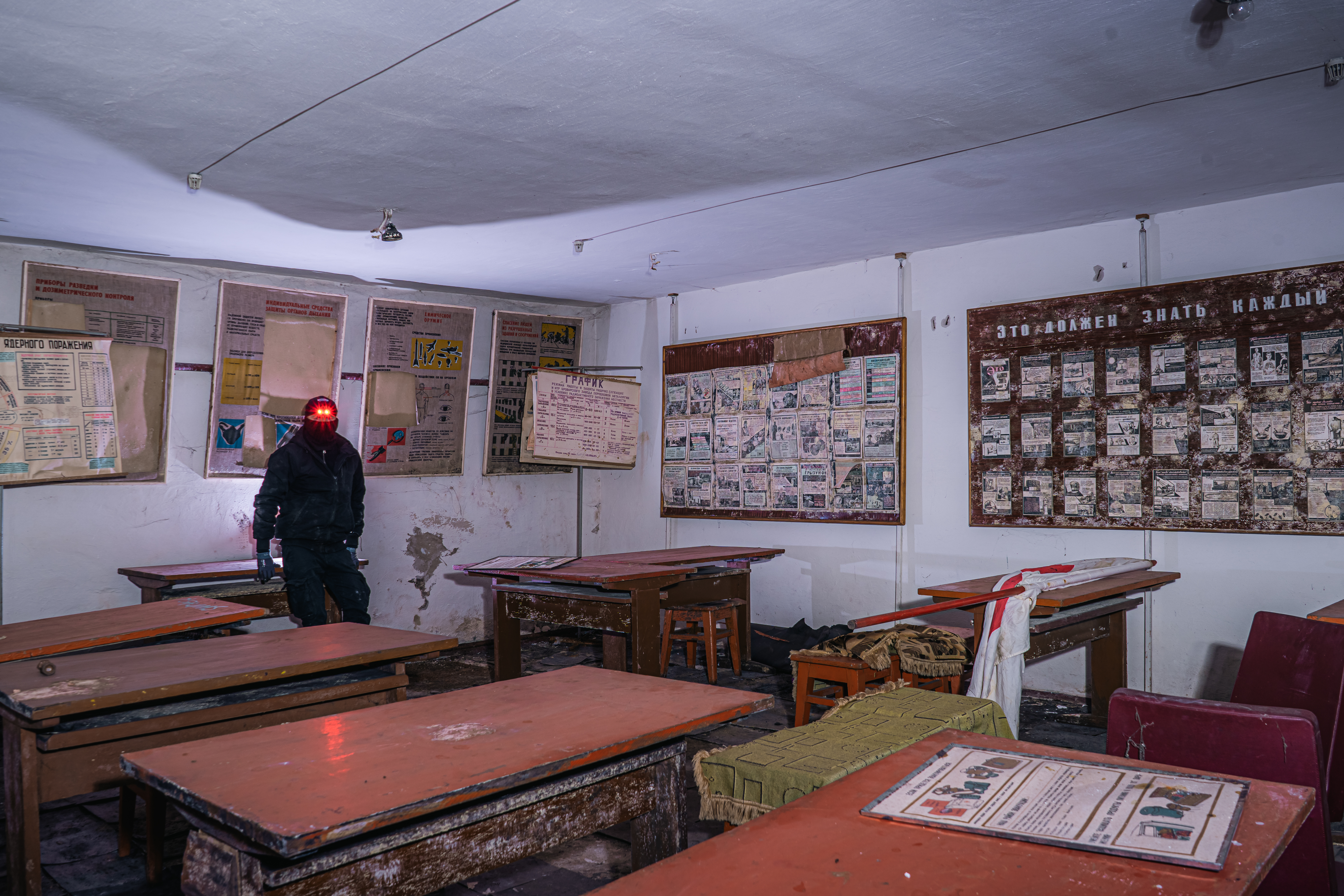 An abandoned classroom with wooden desks and faded educational posters on the walls. A person in dark clothing stands in the corner, their head illuminated by a red light.