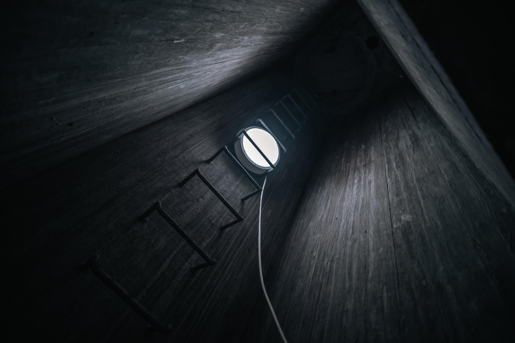 View from the bottom of a dark cylindrical shaft with a metal ladder and a bright circular opening at the top.