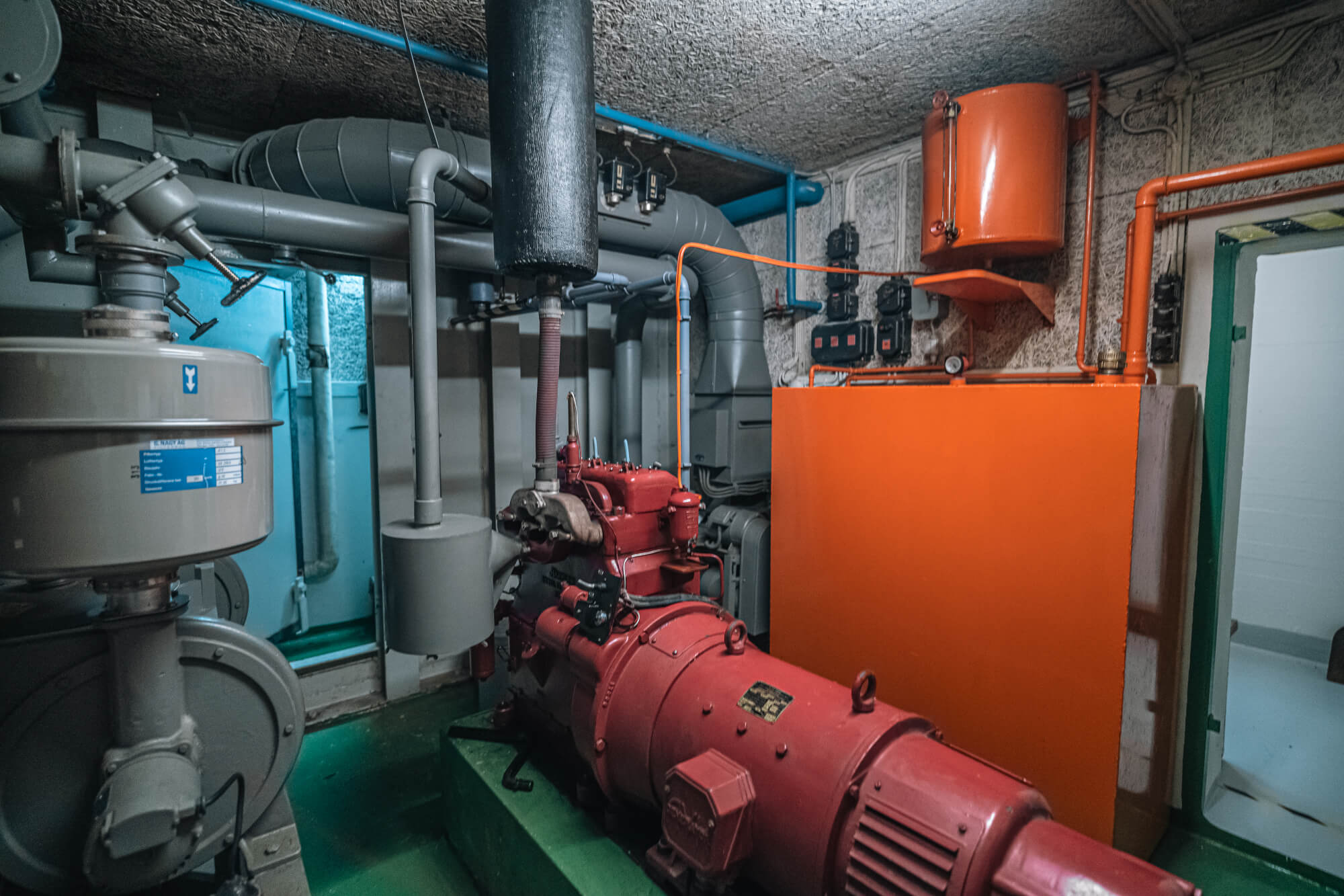 A view of a mechanical pump room featuring various pipes and machinery. A prominent red pump is centered in the scene, surrounded by grey piping and an orange panel on the wall. The room has teal accents and low-lighting, contributing to a utilitarian atmosphere.