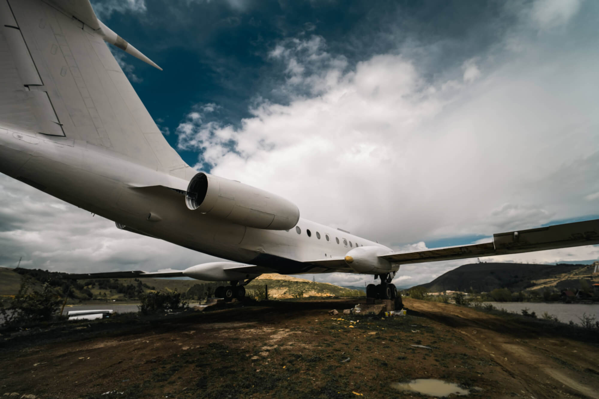 A large white jet airplane positioned on a muddy ground under a cloudy sky, showcasing its rear and wing with a rugged landscape in the background.