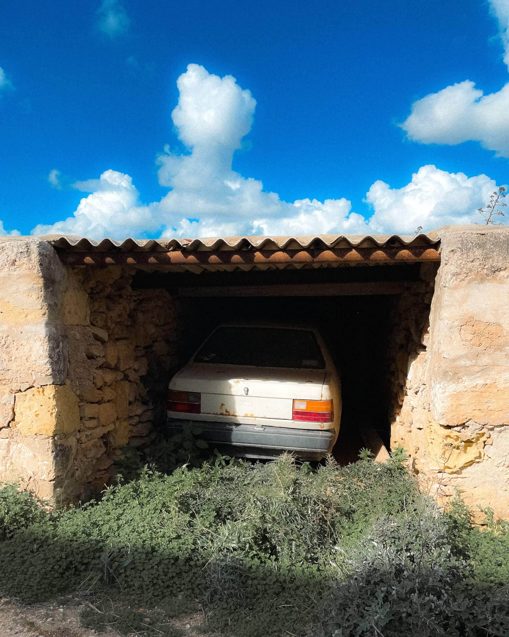 A white car partially visible in a dim stone garage, surrounded by tall green weeds and under a clear blue sky with fluffy white clouds.