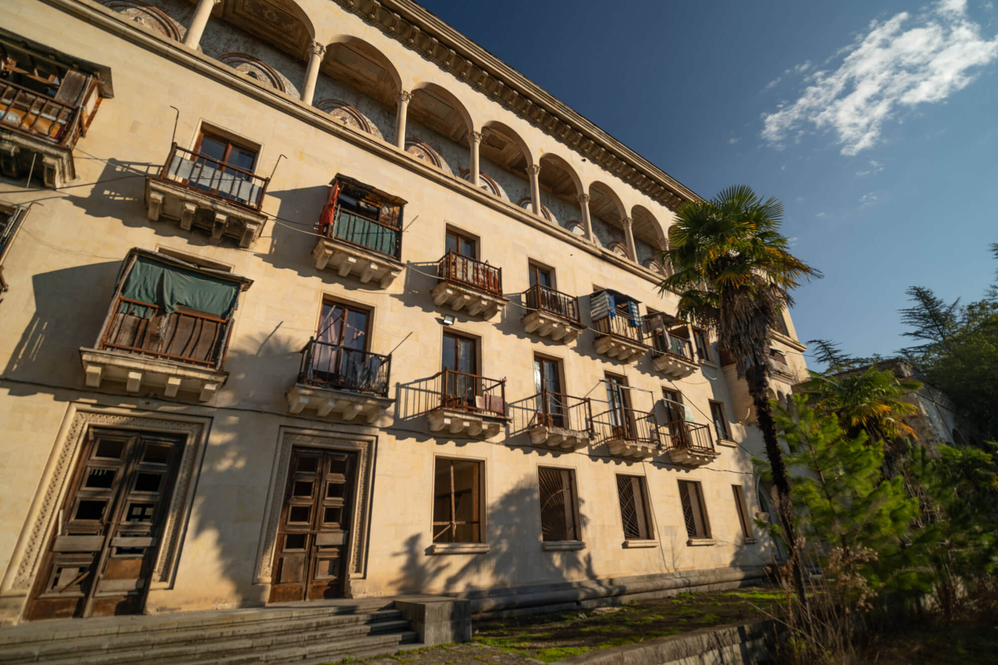 A tilted view of a faded building featuring several balconies with hanging fabrics, wooden doors, and patches of greenery in the foreground under a clear blue sky.