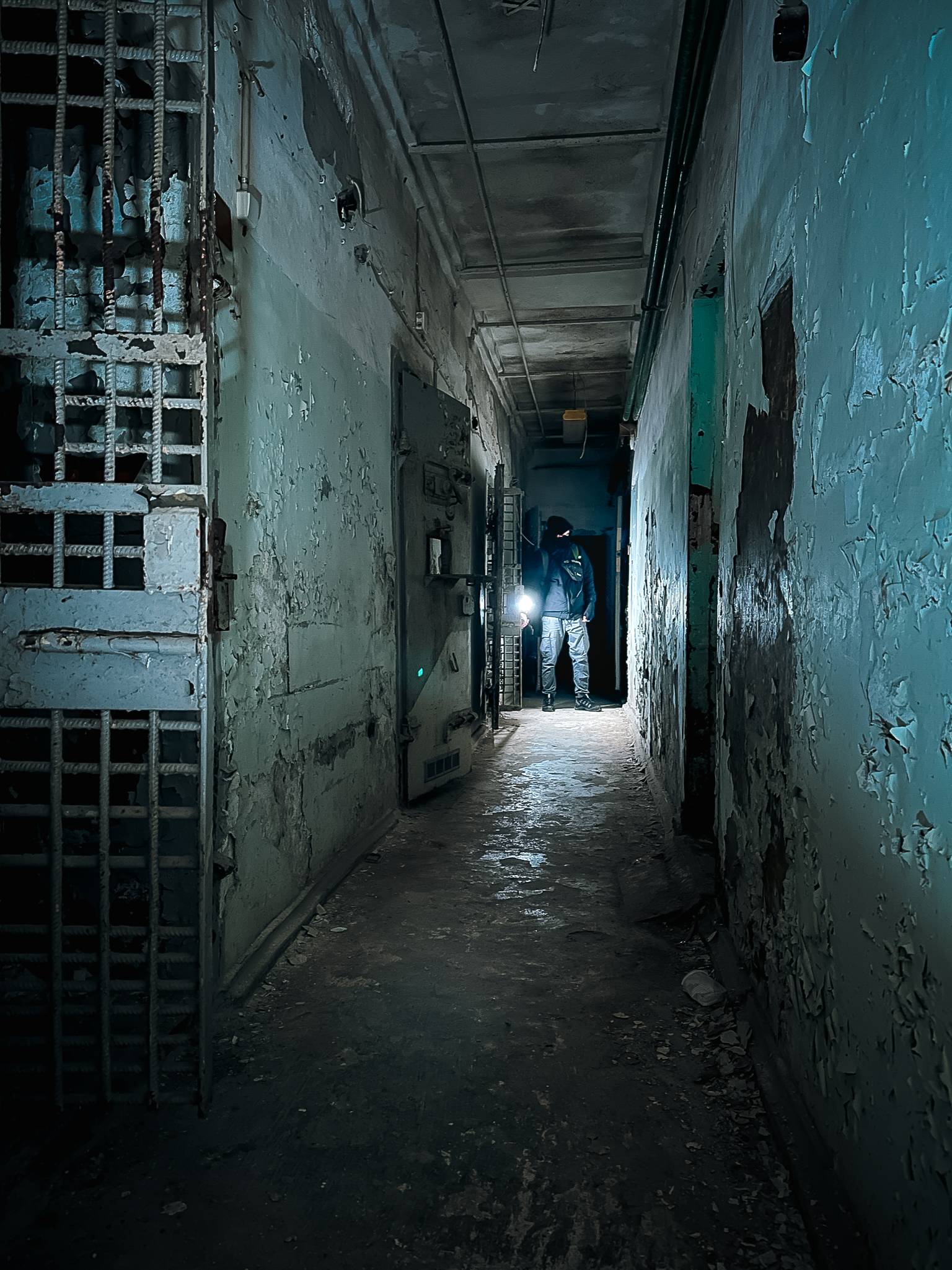 A narrow corridor inside an abandoned building with peeling paint on the walls. To the left, there is a locked prison cell with steel bars, while a figure stands at the end of the hall, illuminated by a flashlight.