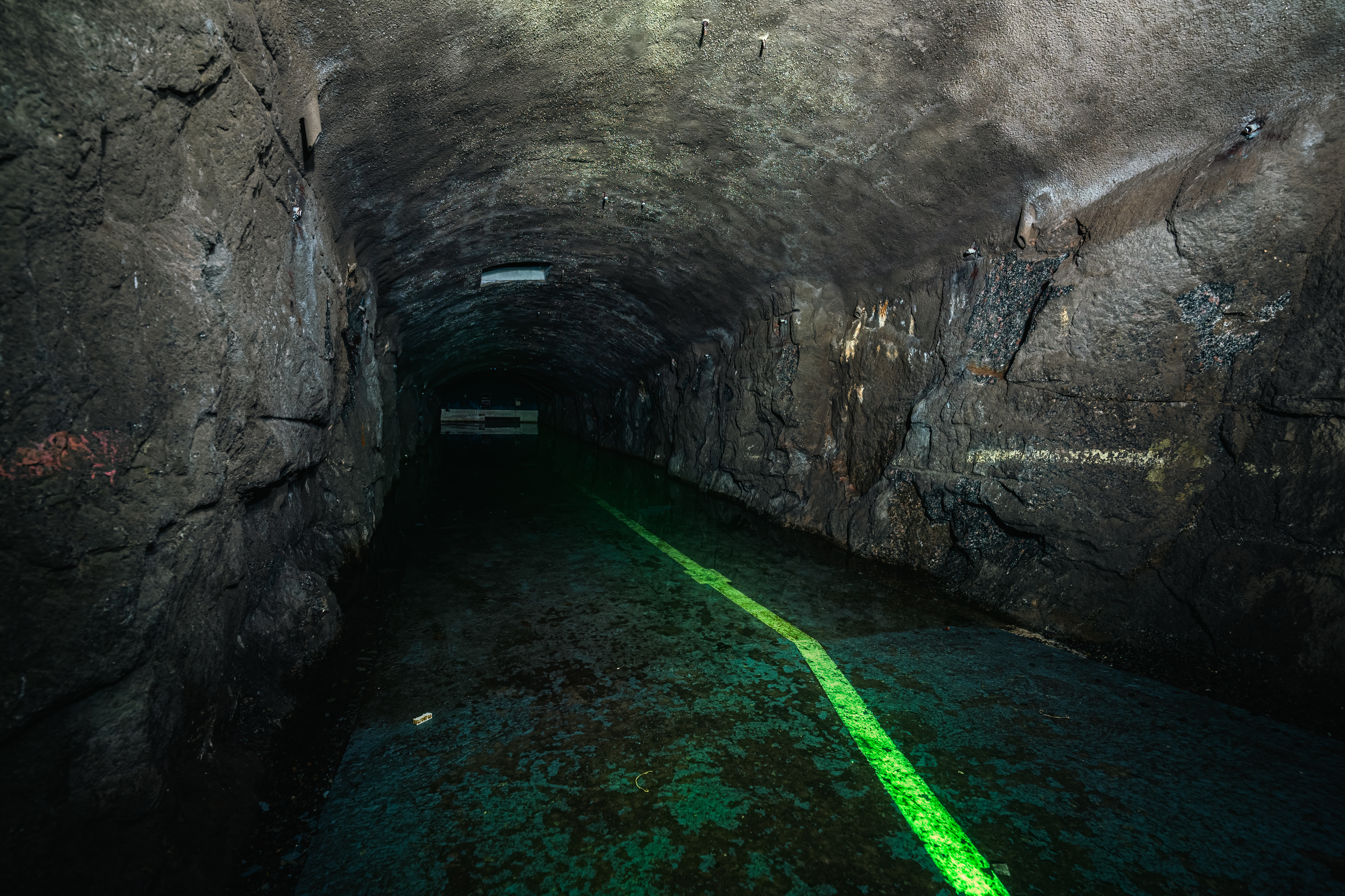 An underground tunnel with rough stone walls and a wet floor, featuring a bright green line marking the path. The space is dimly lit, creating a mysterious and eerie atmosphere.