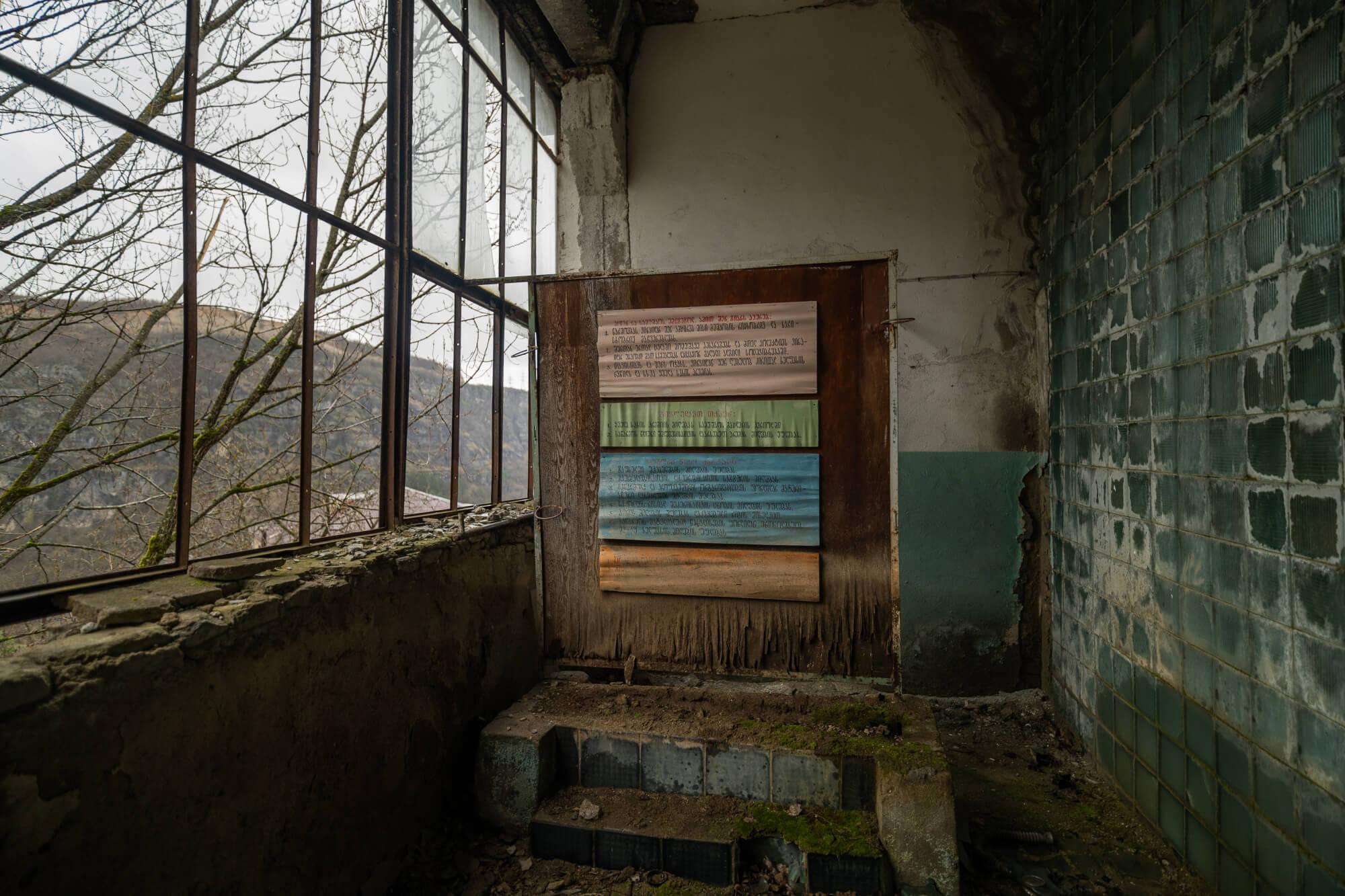 Interior of an abandoned building with a large window showing bare trees and hills outside. A wooden panel with painted text in different colors is mounted on a wall. The floor shows moss and debris, creating an atmosphere of decay.