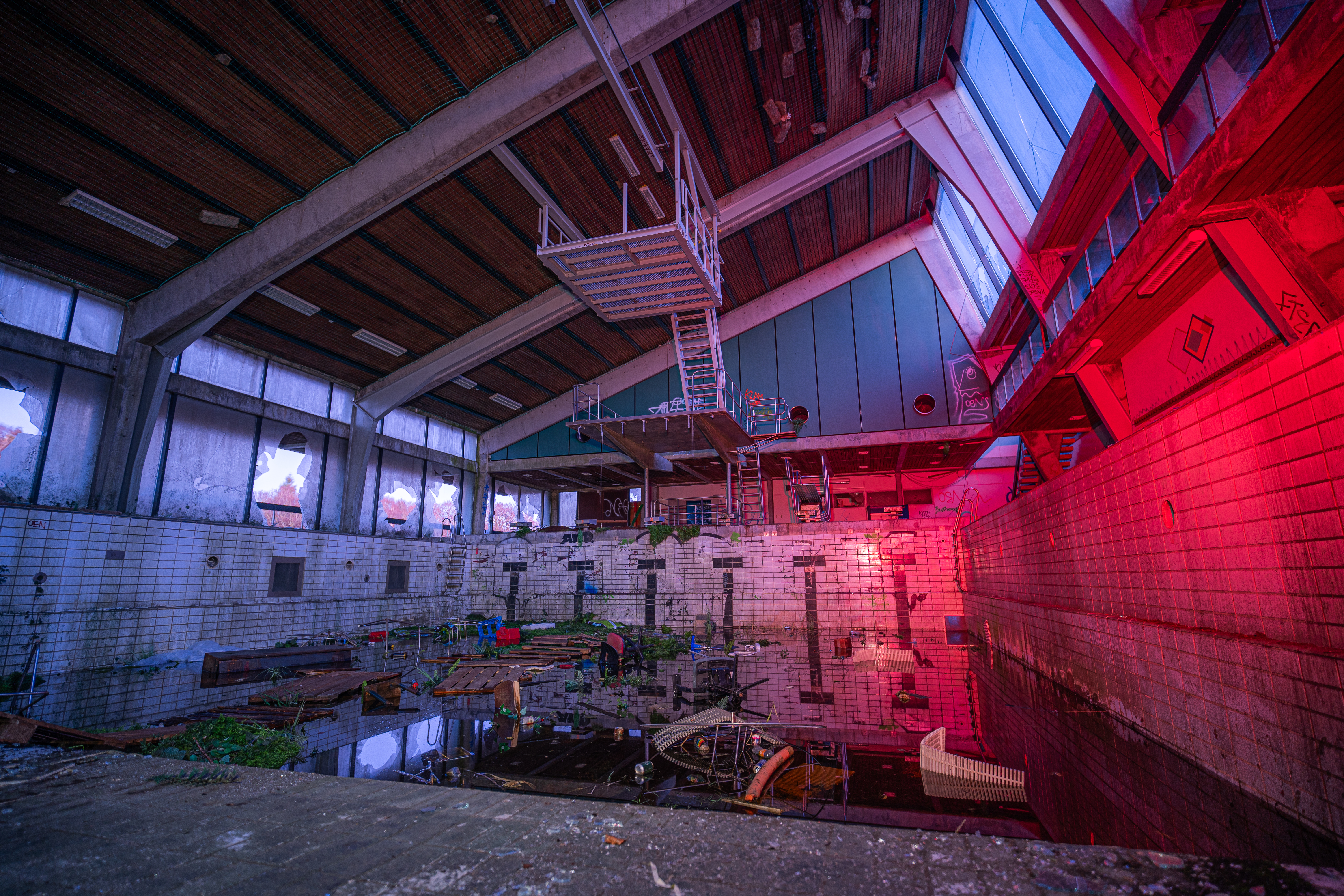 An abandoned indoor swimming pool with broken windows, scattered debris, and water reflecting the remnants of the space. The environment is illuminated by a blend of natural light and artificial reds, creating a haunting atmosphere.