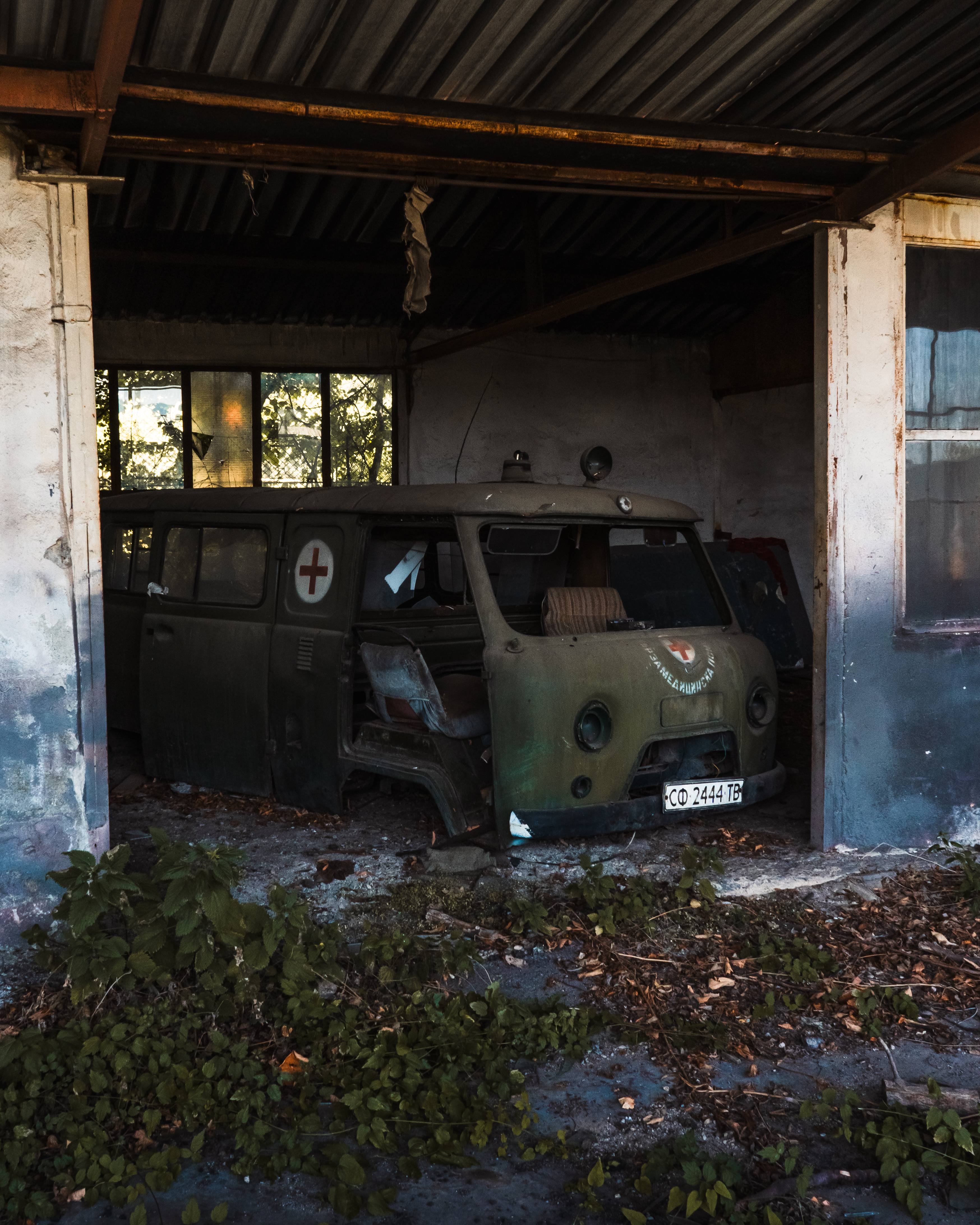 An abandoned green medical vehicle with a red cross emblem, partially visible in a dilapidated building. Surrounding it is overgrown foliage and debris on a concrete floor, with sunlight filtering through dirty windows.
