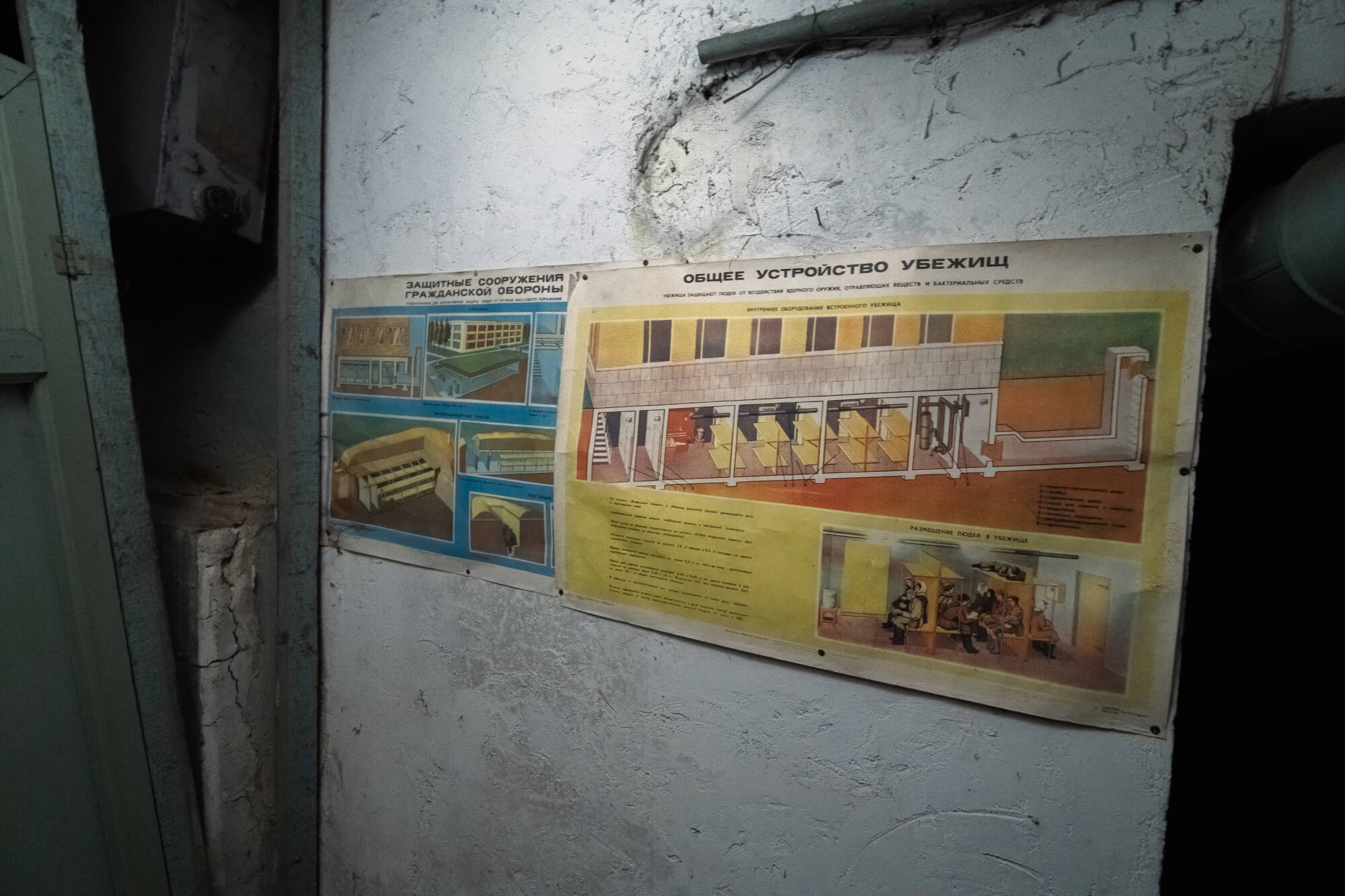 A faded poster on a wall showing diagrams of civil defense structures, with a dimly lit corridor and cracked walls surrounding it.
