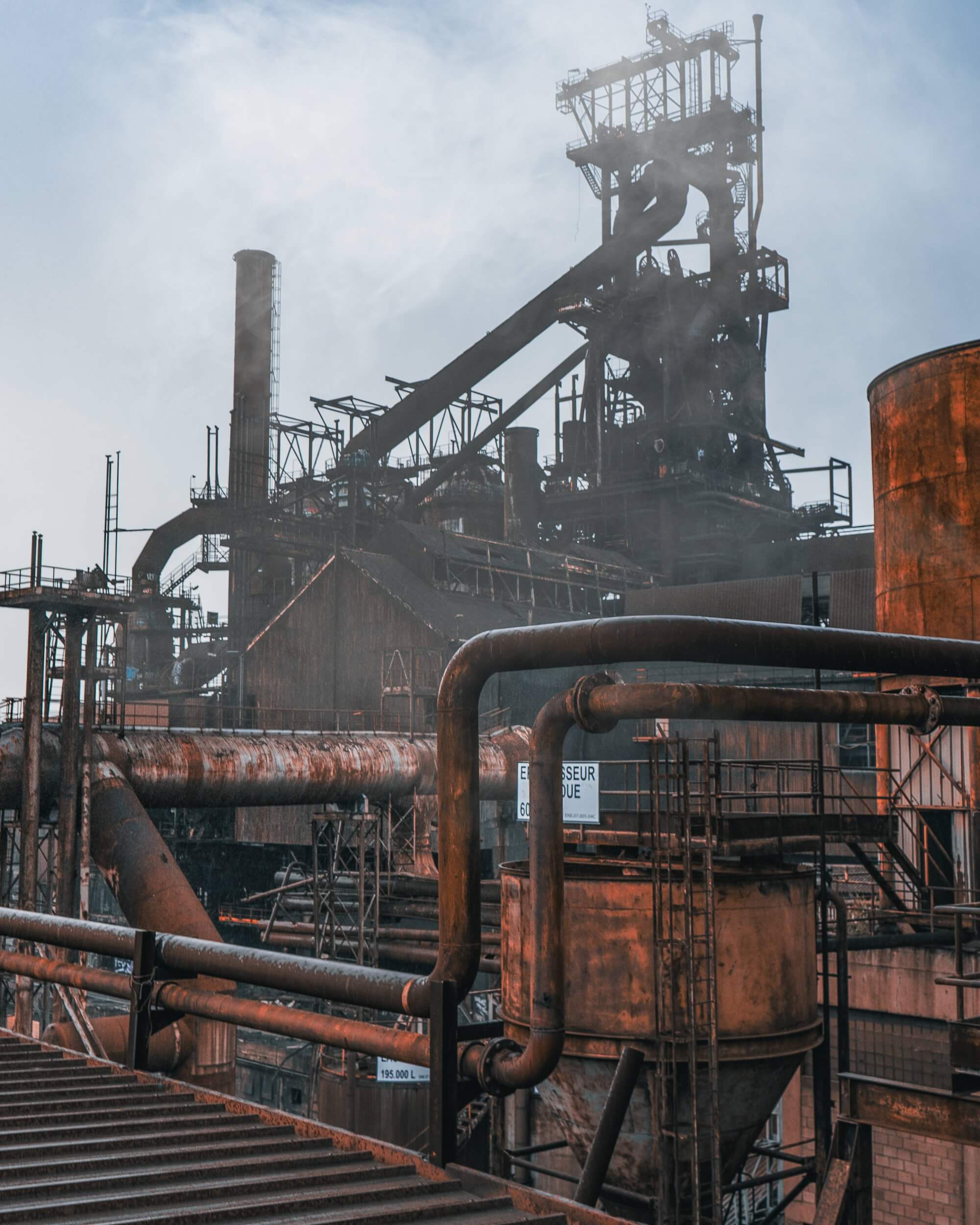 A large industrial complex with smokestacks and metal structures in a misty atmosphere. The foreground features rusted pipes and metal fittings, conveying a sense of scale and complexity.