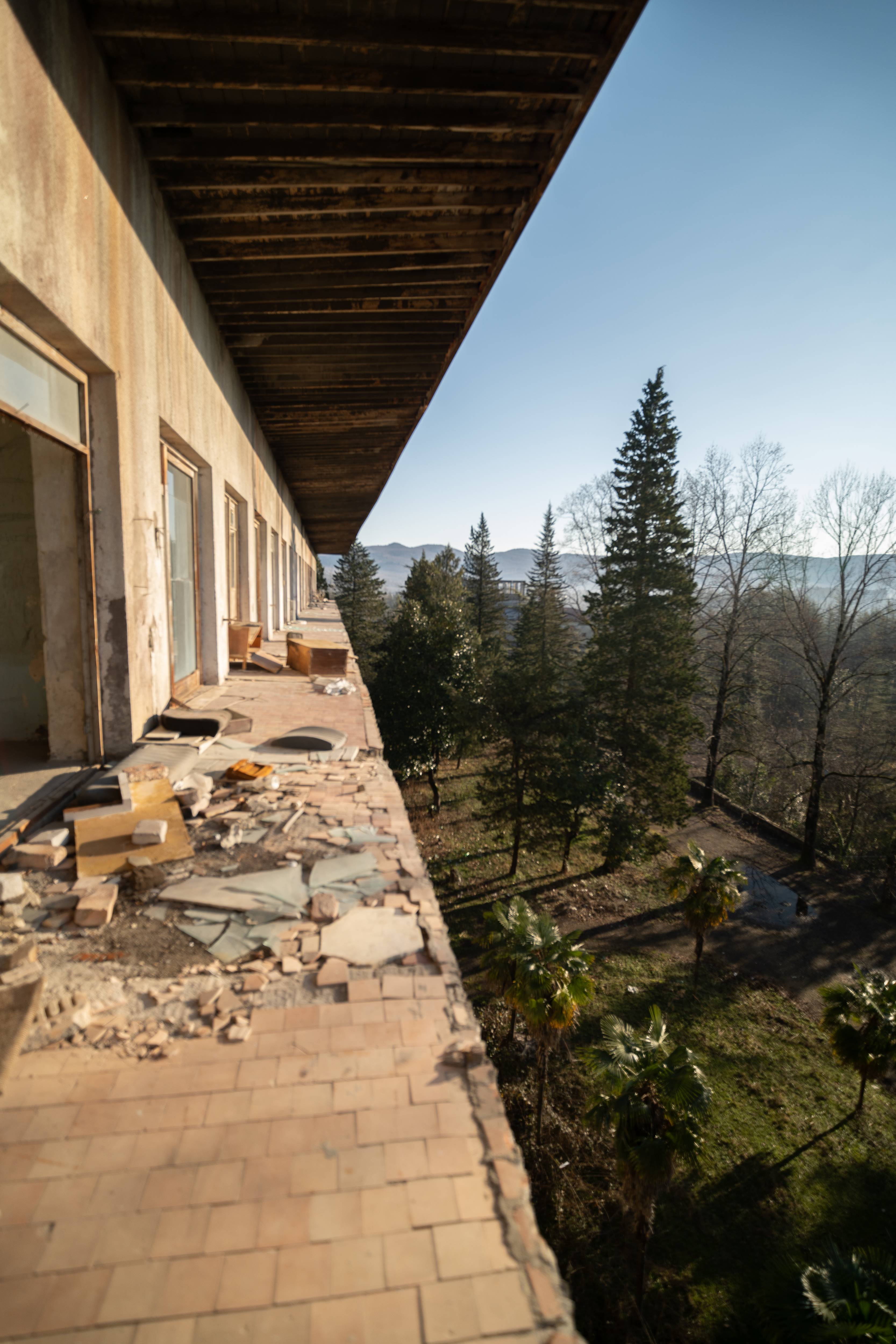 A weathered balcony with broken tiles and debris, overlooking a grove of trees and mountains in the distance under a clear sky.