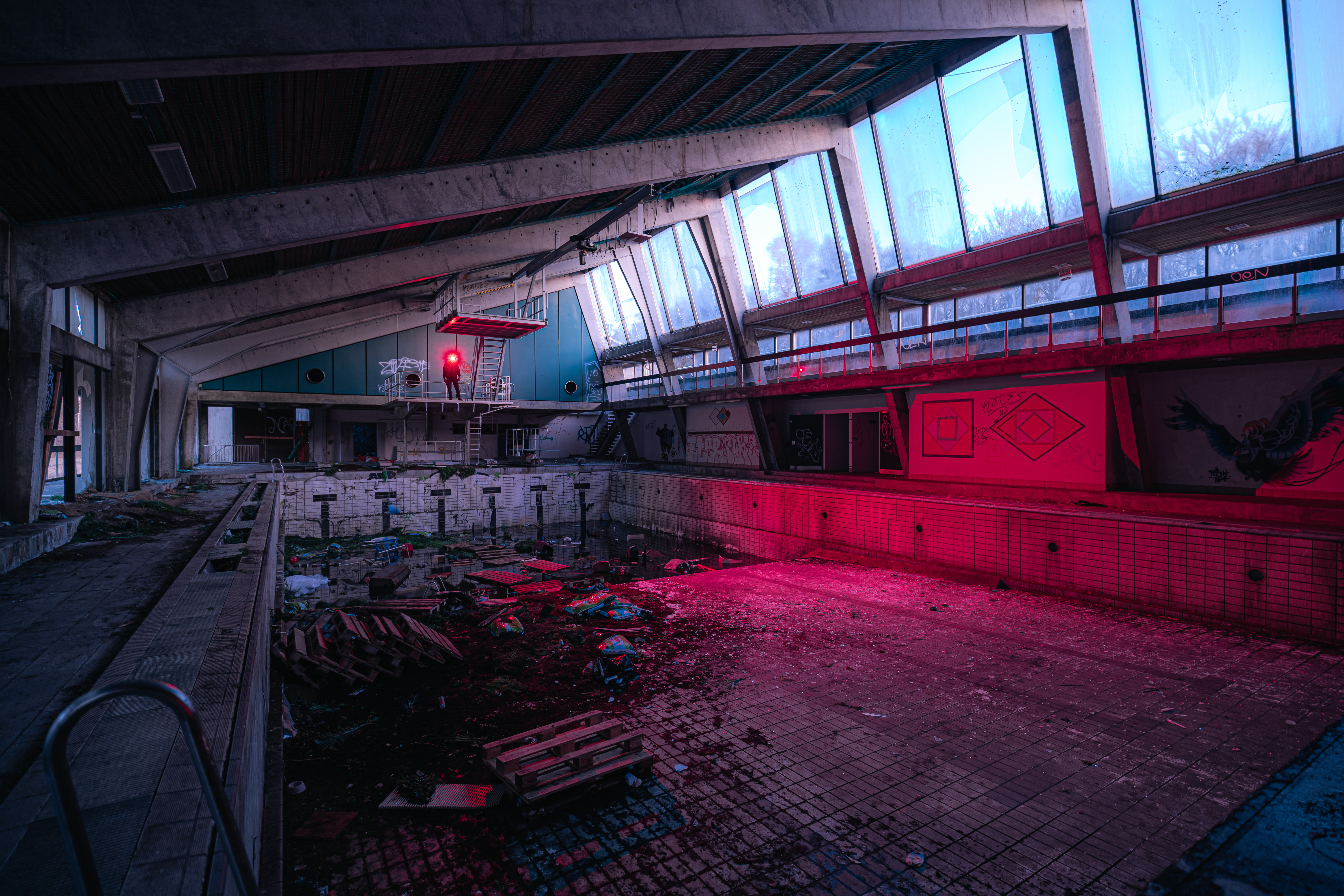 An abandoned indoor swimming pool with a diving board and large windows. The scene is filled with debris, overgrown plants, and graffiti on the walls. A red light illuminates part of the pool area, creating a dramatic contrast with the soft natural light coming through the windows.