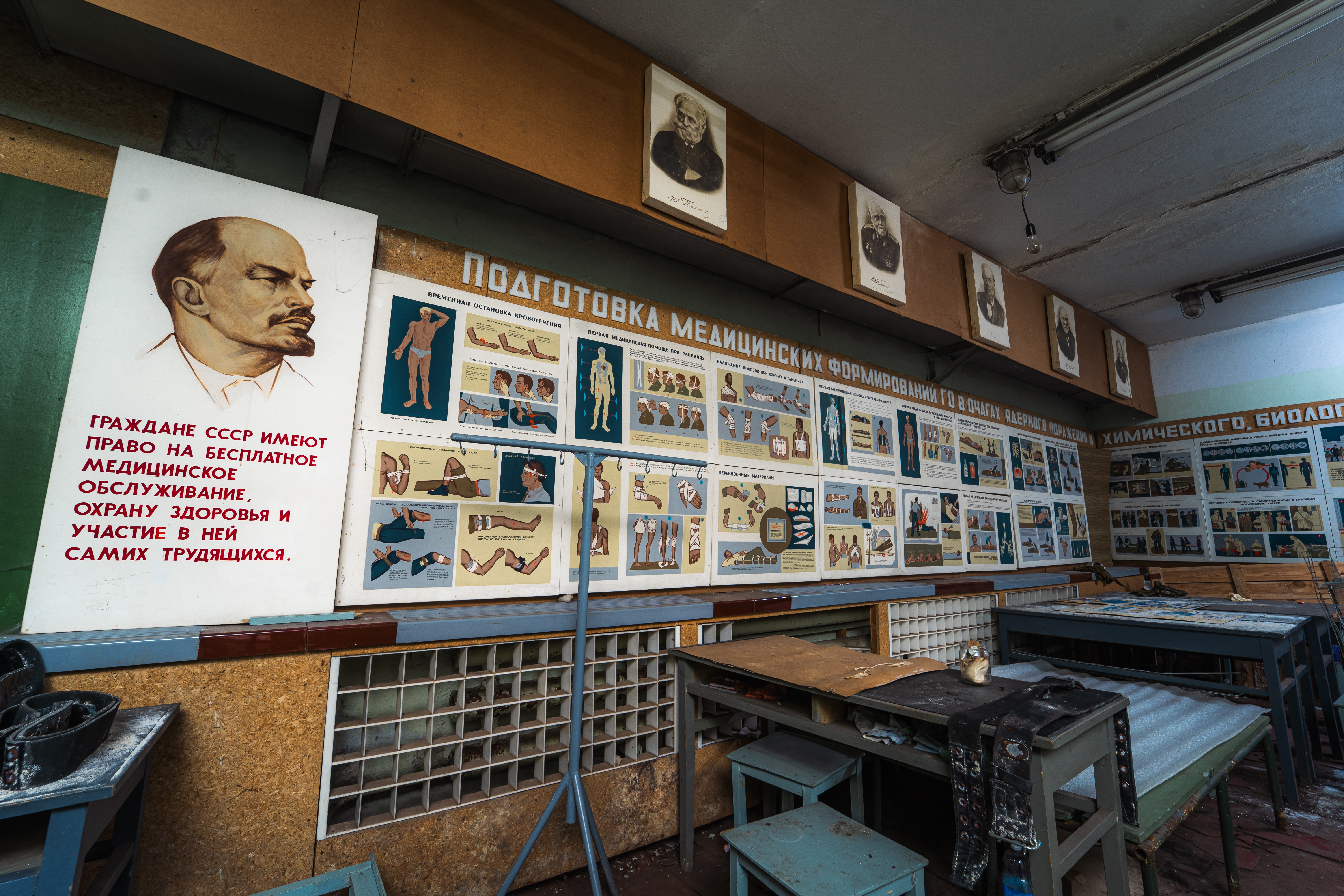 A classroom with vintage medical illustrations on the walls, featuring a portrait of Lenin. There are tables with old tools and instructional charts detailing human anatomy.